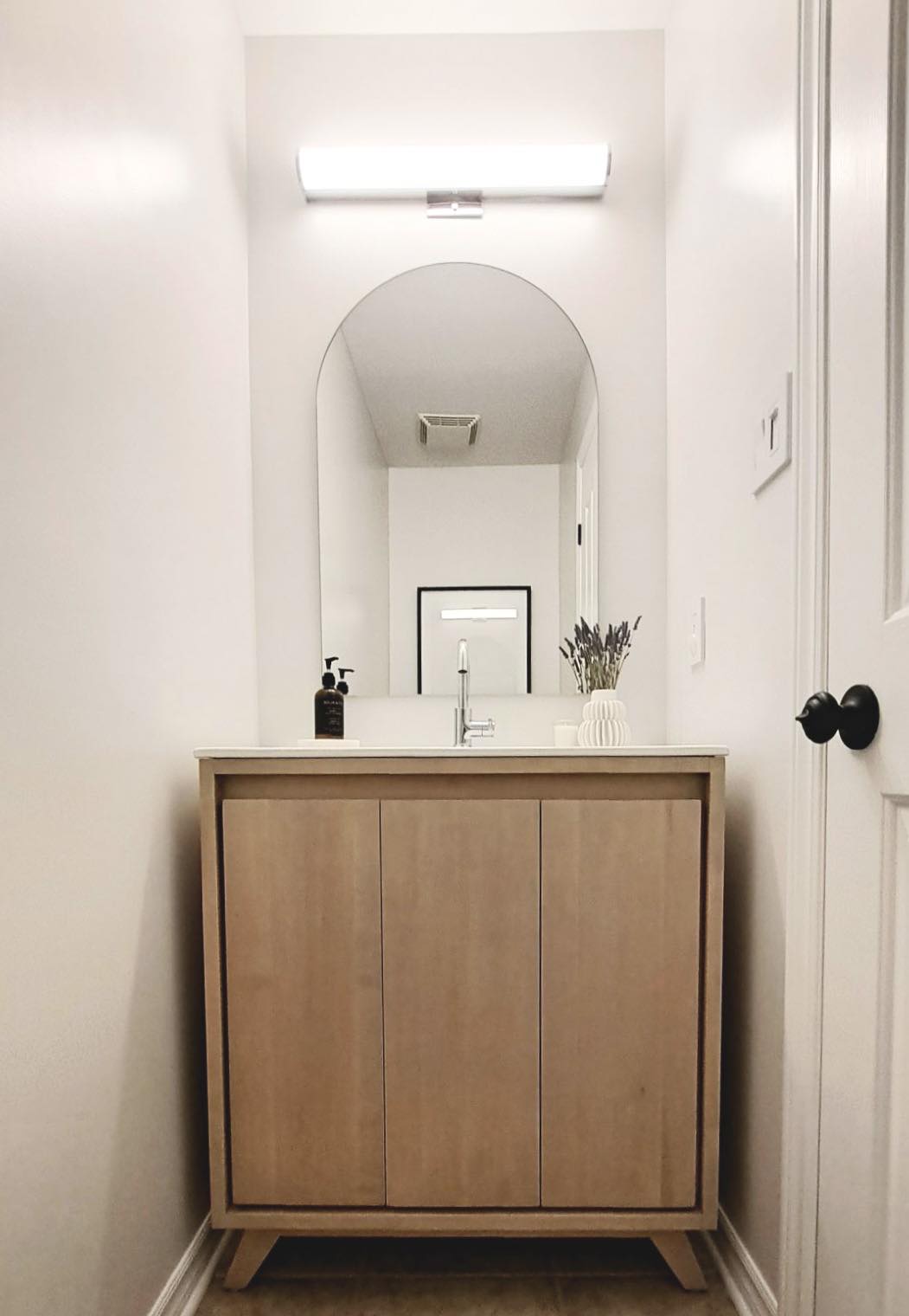 White bathroom walls, light wood cabinet, one-sided rounded mirror, black door handle